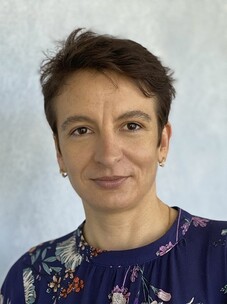 Markova mit kurzem braunem Haar, blauer Bluse mit Muster, vor hellem Hintergrund.